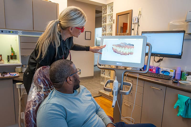 A clinician pointing at a 3D scan of a patient's teeth on a chairside monitor while the patient watches