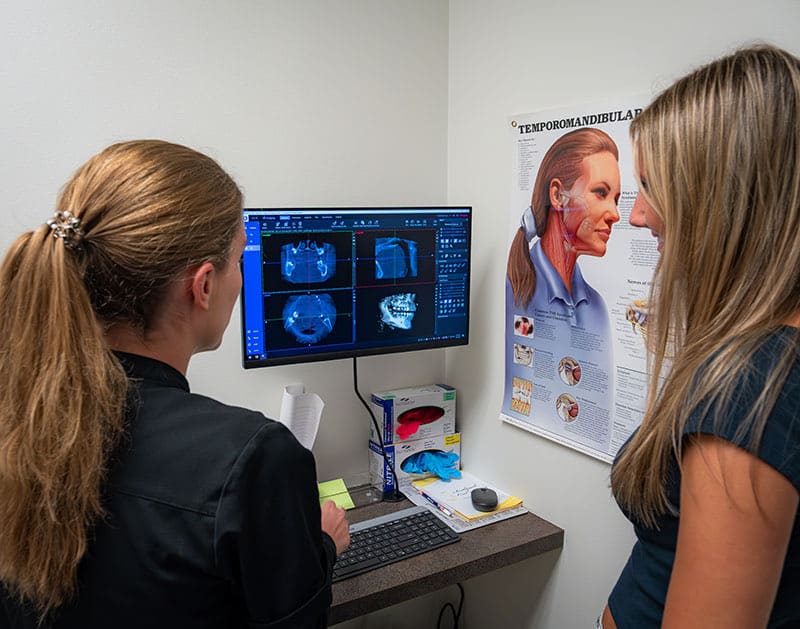 A clinician and a patient looking at 3D jaw-joint scans on a monitor, beside a temporomandibular joint poster